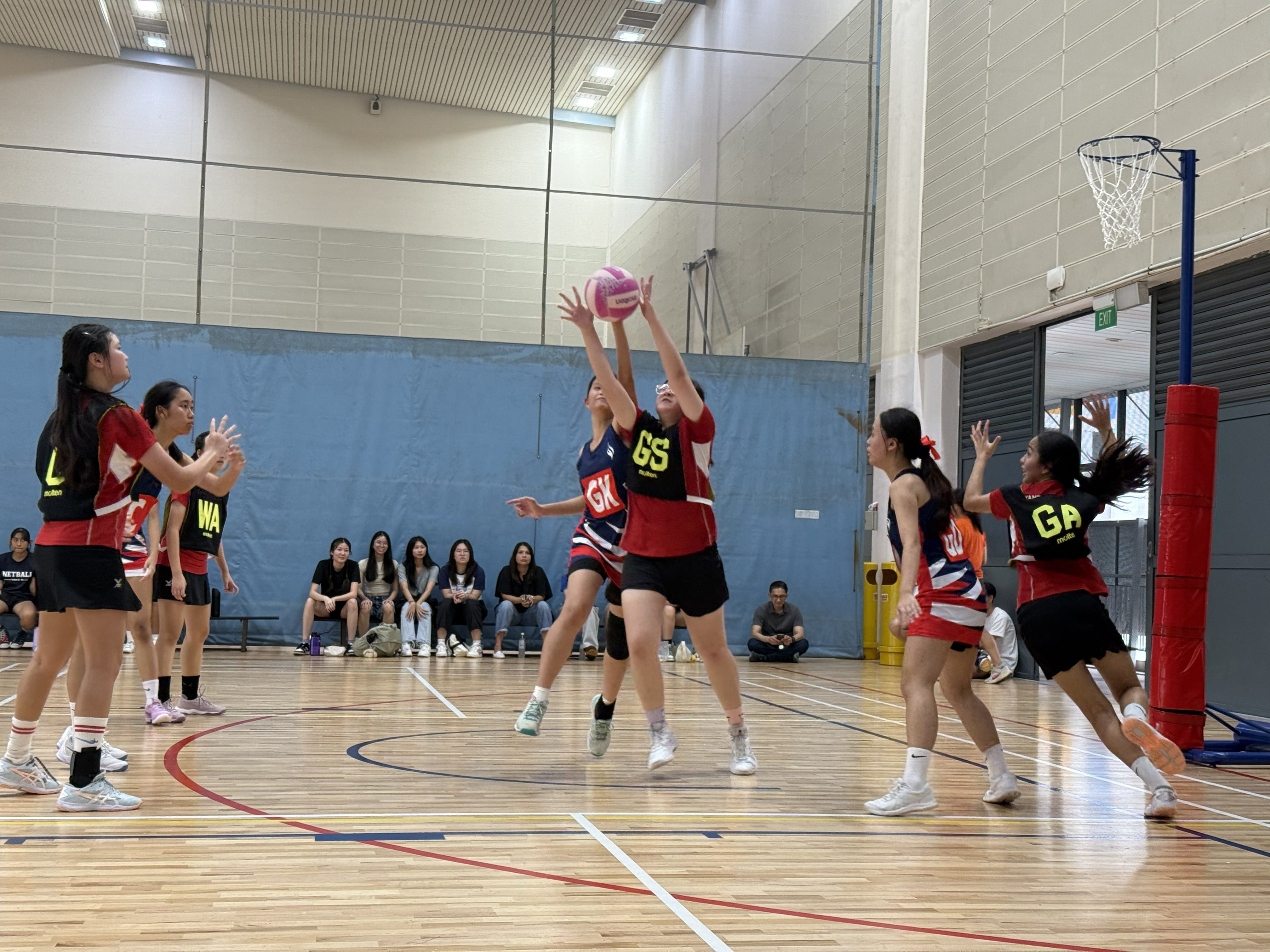 A TMJC player in a red GK bib contests a high ball against an opposing GS player near the goal post during an indoor netball match, with teammates and spectators visible in the background.